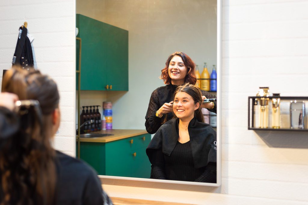 Eine lächelnde Friseurin mit kastanienbraunem Haar stylt das Haar einer lächelnden Kundin mit einem Glätteisen, gesehen durch das Spiegelbild eines großen rechteckigen Spiegels in einem modernen Salon mit smaragdgrünen Schränken.