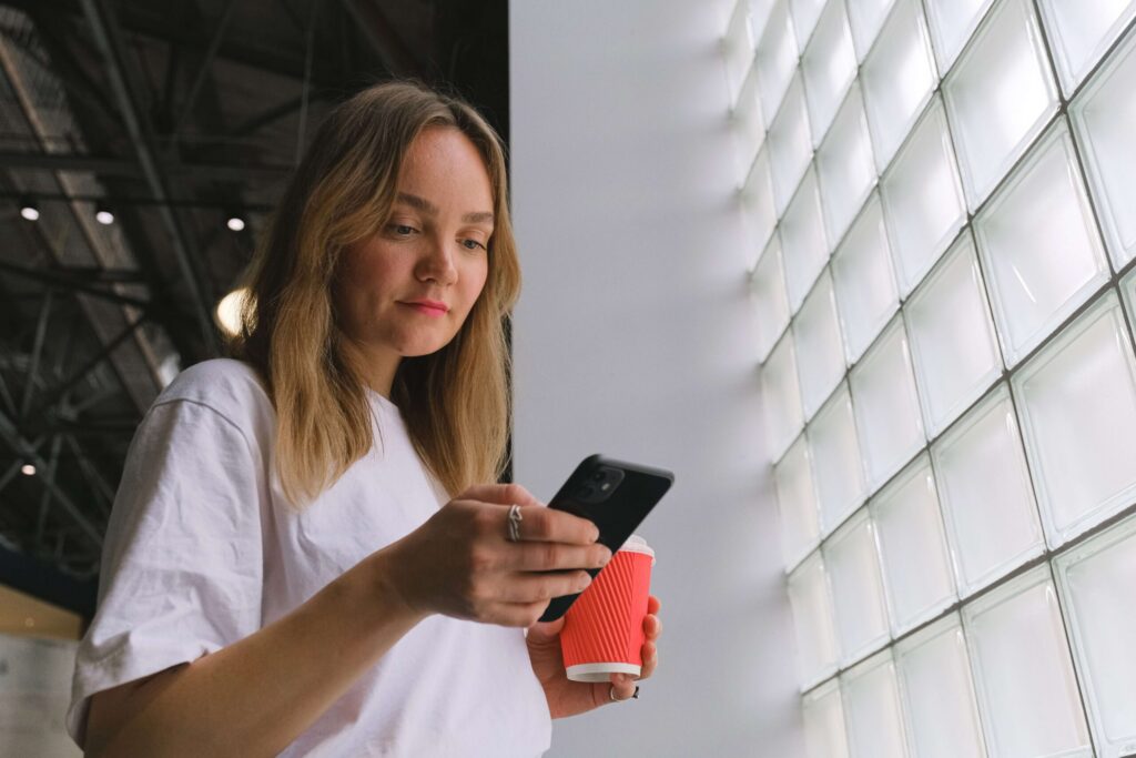 Eine Froschperspektive einer jungen Frau mit blonden Haaren in einem weißen T-Shirt, die auf ihr schwarzes Smartphone blickt, während sie einen roten Kaffeebecher zum Mitnehmen hält. Sie steht neben einer großen Wand aus Glasbausteinen.