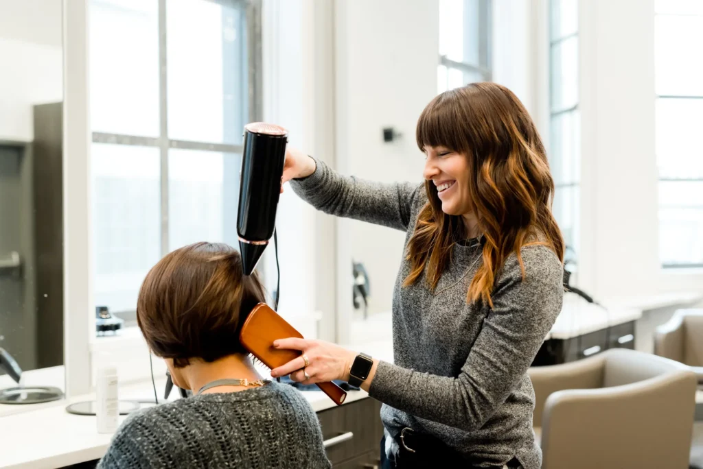 Eine lächelnde Friseurin föhnt und bürstet die Haare eines Kunden in einem hellen, modernen Salon.