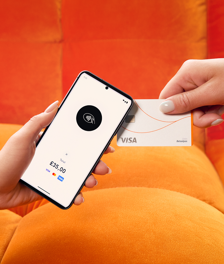 Eine Hand hält ein Smartphone, das als Kartenlesegerät fungiert und ein Symbol für kontaktloses Bezahlen sowie einen Gesamtbetrag von 35,00 £ anzeigt, während eine andere Hand eine weiße Visa-Kreditkarte zum Bezahlen heranhält. Der Hintergrund ist ein leuchtend orangefarbenes Sofa.